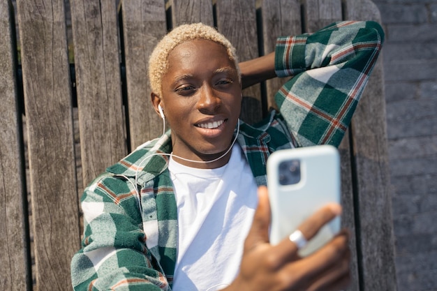 Ragazzo felice con l'acconciatura alla moda che usa musica d'ascolto del telefono cellulare prendendo selfie per strada