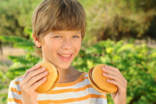 Ragazzo felice dell'adolescente che mangia hamburger all'aperto sopra lo sfondo della natura. Cibo spazzatura malsano