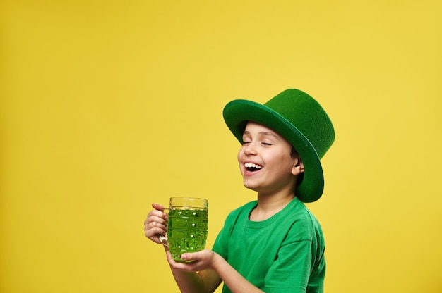 Ragazzo felice in cappello del leprechaun che gode della bevanda verde che sta sul backgroud giallo con lo spazio della copia