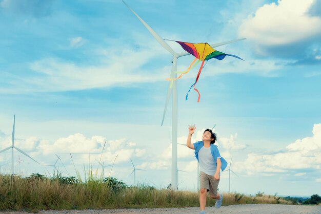 Ragazzo felice progressivo che corre e fa volare l'aquilone con il fondo della turbina eolica