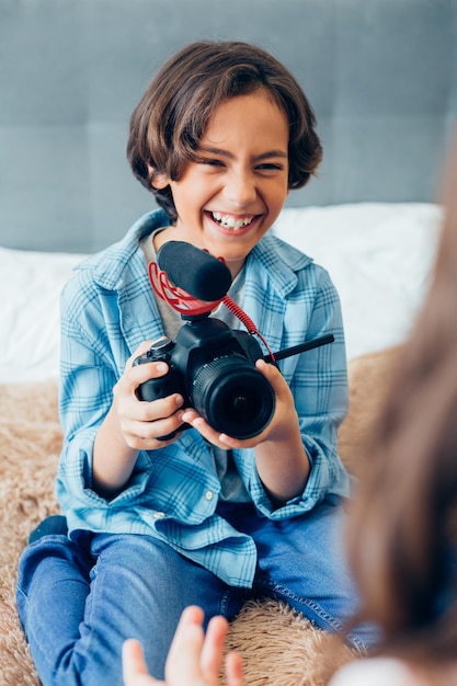 Ragazzo felice seduto su un letto a casa con una moderna videocamera a casa e ridendo