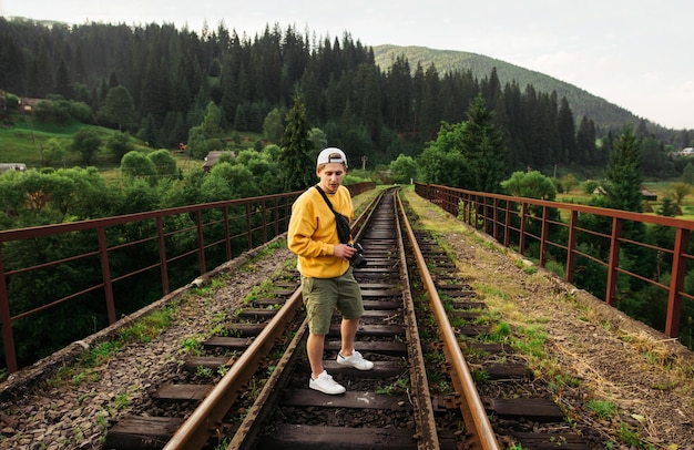 Ragazzo fotografo con una macchina fotografica in mano si trova su un ponte ferroviario in montagna