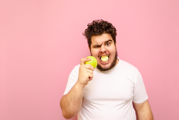Ragazzo grasso divertente in maglietta bianca che mangia una mela verde ed esamina la macchina fotografica con una faccia seria