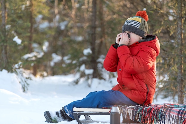 Ragazzo in abiti da inverno seduto su una slitta e che si copre la faccia con le mani e beve tè