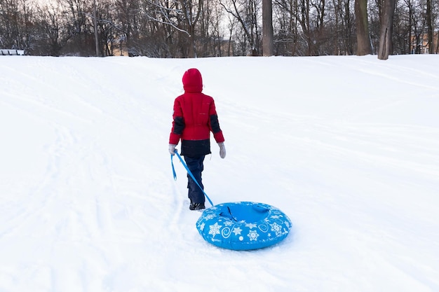 Ragazzo in abiti invernali che cammina con tubi blu tra neve bianca, vista posteriore. Gite in slitta gonfiabile.