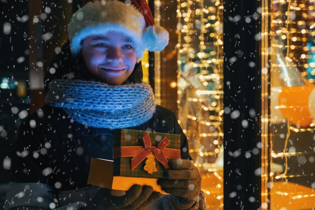 Ragazzo in cappello di Babbo Natale con scatola regalo vicino alla vetrina illuminata. Natale presenta le vacanze o lo shopping per il nuovo anno o il concetto di Natale