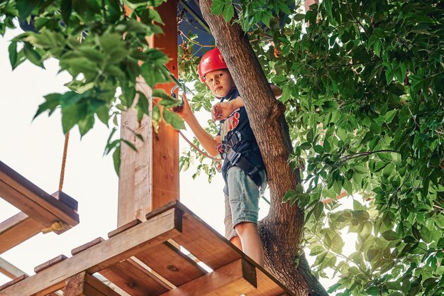 Ragazzo in casco e attrezzatura da arrampicata in piedi sulla piattaforma sullo sfondo del fogliame nel parco delle funi