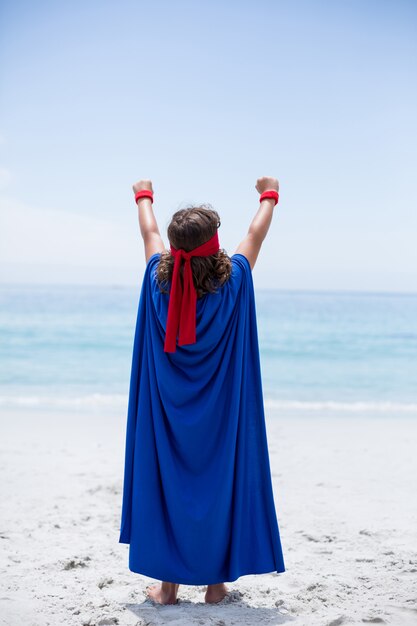 Ragazzo in costume da supereroe con le braccia alzate in spiaggia