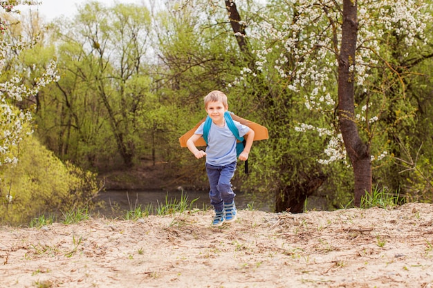 Ragazzo in età prescolare allegro che corre vicino al fiume con ali di legno sulla schiena Il ragazzino creativo usa lo zaino per sistemare le ali di legno sulla schiena Sognando di essere un pilota Creatività e interesse per bambini