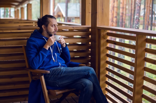 Ragazzo in giacca blu con un cappuccio drappeggiato sulla testa seduto su una sedia in balcone con il tè in mano