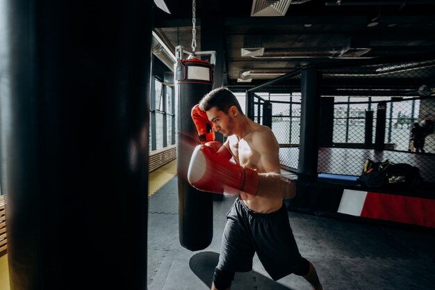 Ragazzo in guanti da boxe rossi con un torso nudo vestito con pantaloncini neri colpisce il sacco da boxe in palestra.