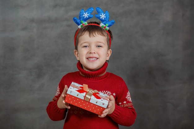 Ragazzo in maglione di Natale e orlo di corno di cervo con piccolo regalo di Natale. Foto di alta qualità