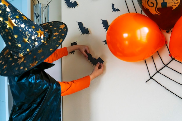 Ragazzo in mantello nero e cappello da strega incolla il pipistrello di carta al muro I bambini decorano la casa per Halloween