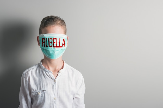 Ragazzo in maschera medica con scritta rossa RUBELLA sul viso su sfondo luminoso.