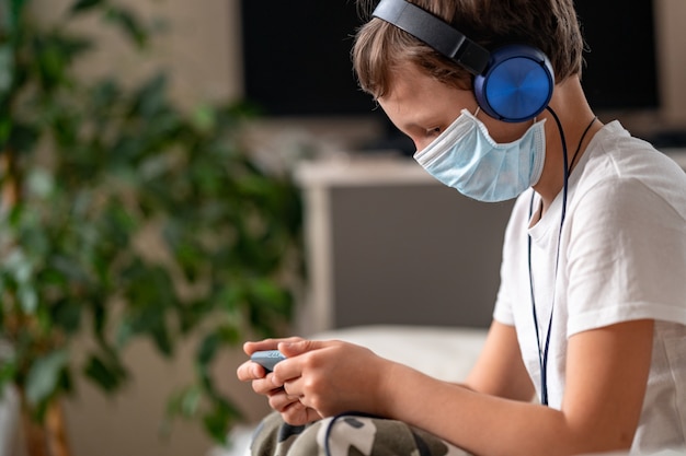 Ragazzo in maschera protettiva e cuffie, con uno smartphone in mano, vista laterale.