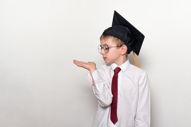 Ragazzo in un cappello accademico quadrato e occhiali regge il palmo della mano. Concetto di scuola. Sfondo bianco.