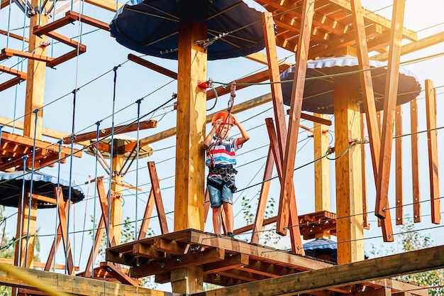 Ragazzo in un casco e attrezzatura da arrampicata in piedi su una piattaforma di legno nel parco delle funi in serata estiva