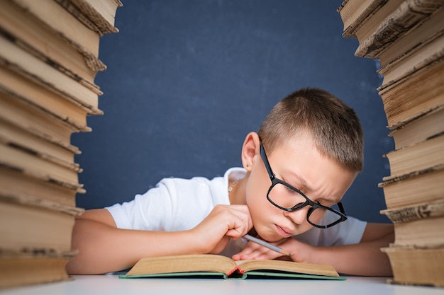 Ragazzo intelligente con gli occhiali seduto tra due pile di libri e leggere il libro pensieroso.