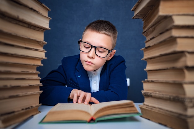 Ragazzo intelligente con gli occhiali seduto tra due pile di libri e leggere il libro pensieroso.