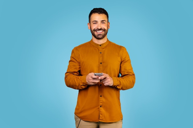 Ragazzo italiano gioioso che tiene uno smartphone moderno che guarda lo schermo della fotocamera su sfondo blu per studio