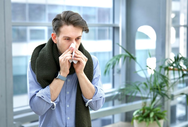 Ragazzo malato malato in sciarpa che si soffia il naso nel fazzoletto di carta giovane con influenza nasale che cola