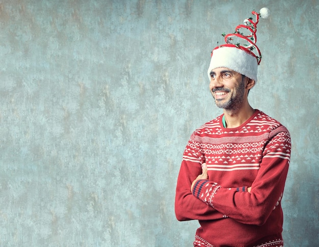 Ragazzo marrone con le braccia incrociate con la barba corta grigia che sorride con un maglione rosso di Natale e un divertente cappello di Natale