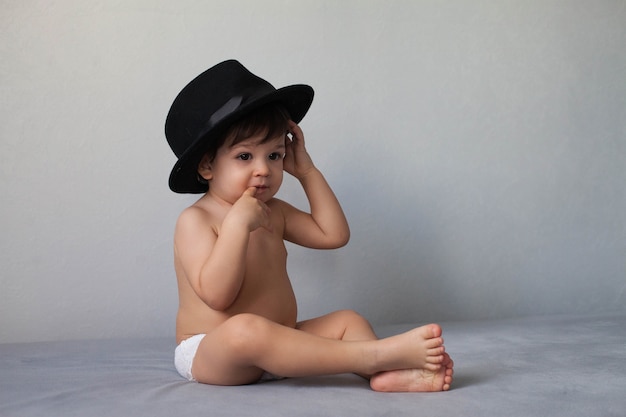 Ragazzo nudo del bambino che indossa un cappello nero e seduto su uno sfondo grigio neutro e con un dito in bocca.