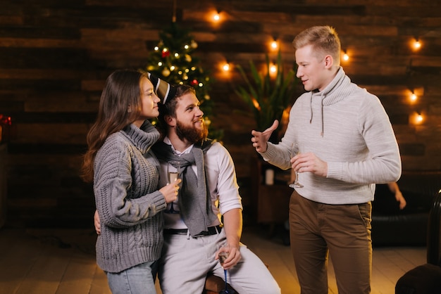 Ragazzo parla con una coppia per celebrare la festa di capodanno in una casa decorata a festa con in mano bicchieri di champagne. Albero di Natale con ghirlanda e illuminazione festiva sullo sfondo.