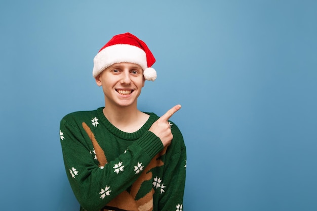 Ragazzo positivo in maglione natalizio e cappello santa si erge su uno sfondo blu guarda a porte chiuse