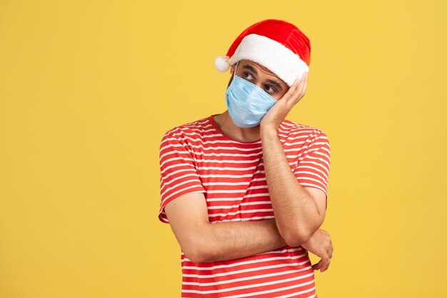 Ragazzo premuroso incerto con cappello da Babbo Natale con maschera protettiva medica che contempla le vacanze trascorse in quarantena mantenendo le distanze Studio indoor girato isolato su sfondo giallo