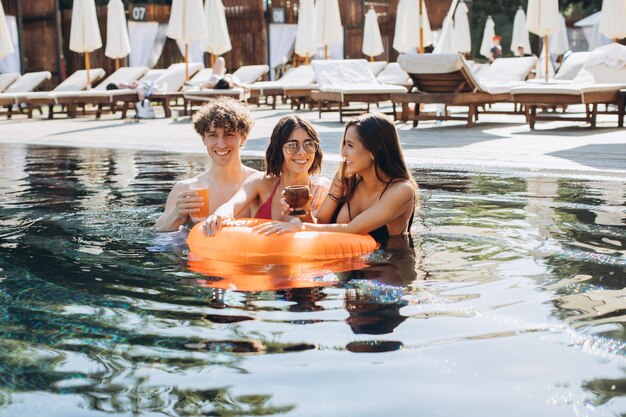 Ragazzo riccio e due belle ragazze che bevono cocktail e si divertono a bordo piscina in estate