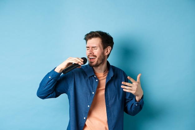 Ragazzo romantico che canta una canzone nel microfono e gesto, cantante che suona il karaoke, in piedi su sfondo blu.