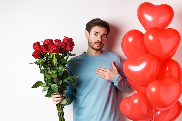 Ragazzo romantico esprimere il suo amore il giorno di San Valentino con regali, portare bouquet di rose rosse e palloncini, tenendo la mano sul cuore, in piedi su bianco