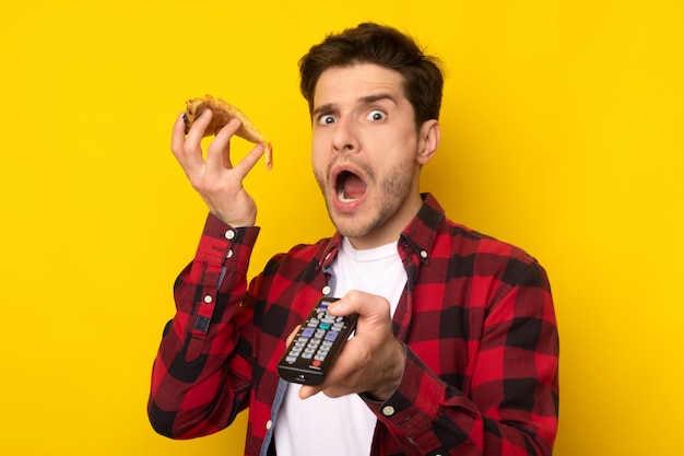 Ragazzo scioccato divertente che mangia pizza tenendo il telecomando guardando la tv