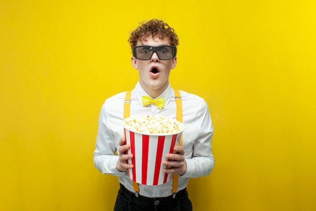 Ragazzo scioccato in abito festivo in occhiali 3d guarda film con popcorn su sfondo giallo isolato