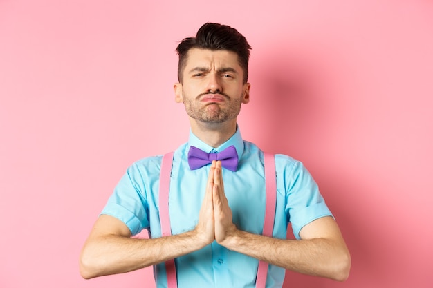 Ragazzo sciocco che fa la faccia implorante, supplica e chiede aiuto, in piedi con le mani premute insieme sul rosa. Copia spazio