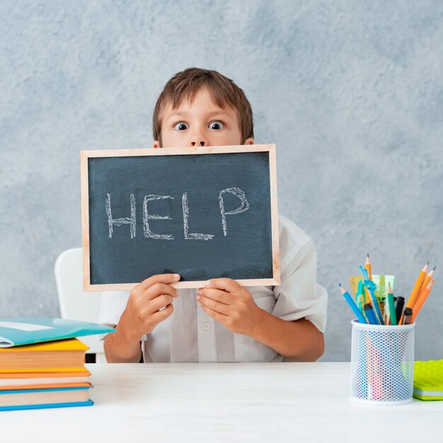 Ragazzo scolaro frustrato stanco triste al tavolo e tenendo la carta con la parola Aiuto. Difficoltà di apprendimento, concetto di educazione