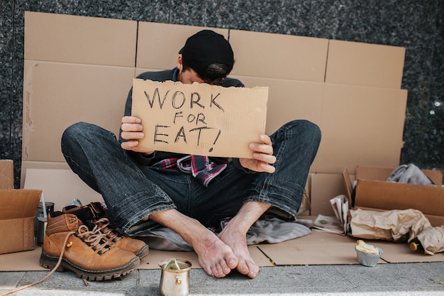 Ragazzo senza lavoro è seduto sul terreno di cemento e si copre il viso con il segno di lavoro per mangiare