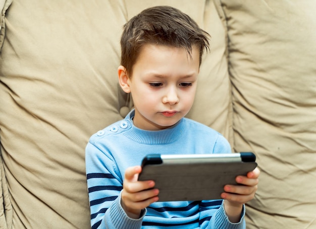 Ragazzo serio che guarda qualcosa di interessante dal tablet digitale seduto su un divano nel soggiorno di casa Gadget e concetto di tecnologia