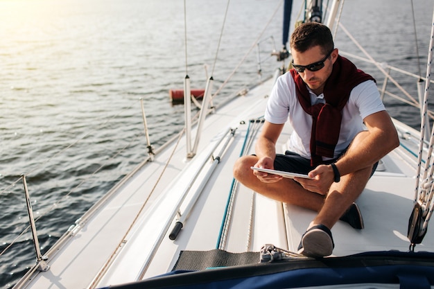 Ragazzo serio e pacifico si siede a bordo dello yacht. Tiene e guarda il tablet. Il giovane è calmo. Indossa una gonna bianca e un maglione scuro con pantaloncini.