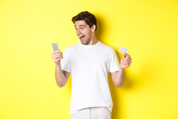 Ragazzo sorpreso con smartphone e carta di credito, shopping online il venerdì nero, in piedi su sfondo giallo.