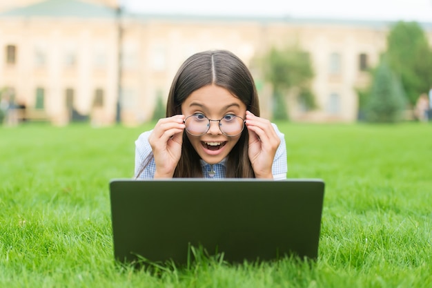 Ragazzo sorpreso della scuola con gli occhiali che gioca online sul computer portatile che si rilassa sull'erba verde nel parco