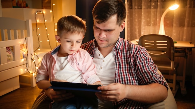 Ragazzo sorridente con il padre che utilizza il computer tablet in camera da letto decorata per il Natale. Concetto di educazione dei bambini e famiglia che hanno tempo insieme di notte.