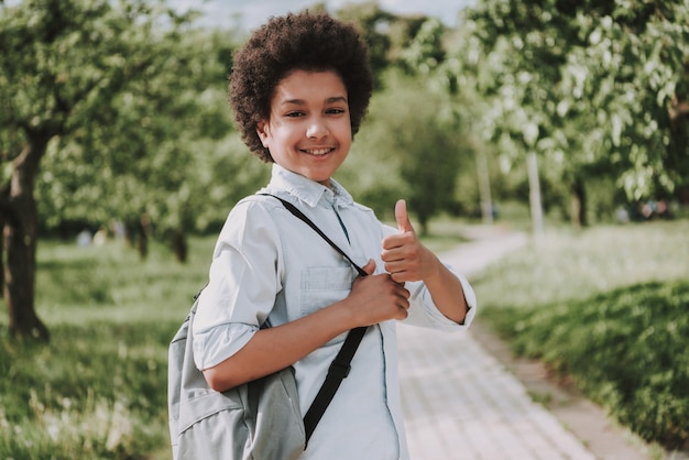 Ragazzo sorridente con zaino Visualizza i pollici in su nel parco.