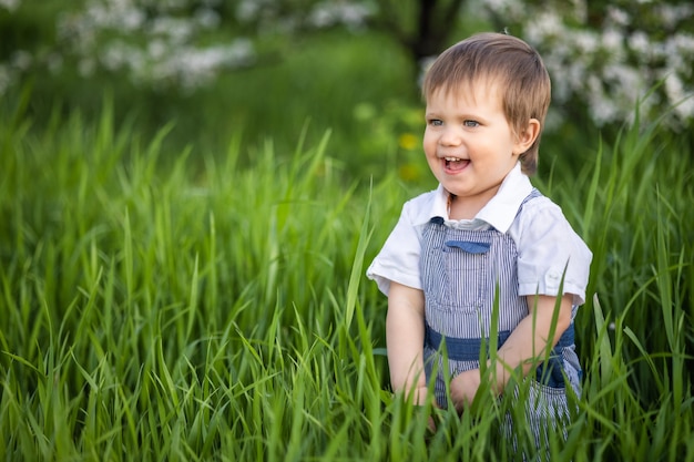 Ragazzo sorridente divertente in tuta blu denim e occhi azzurri luminosi. È divertente nascondersi nell'erba alta e verde in un caldo giardino primaverile sullo sfondo di alberi in fiore.