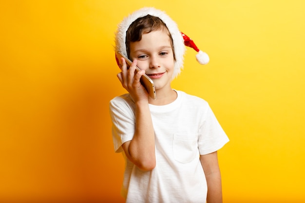 Ragazzo sorridente felice in cappello rosso di Natale con il telefono su colore giallo