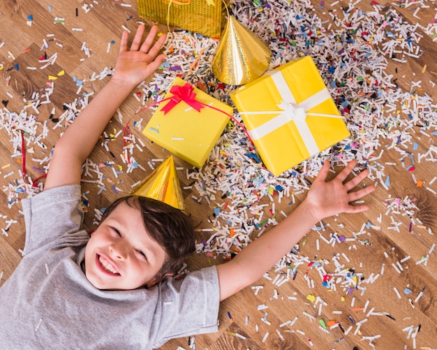 Ragazzo sorridente in cappello di compleanno sdraiato con regali e coriandoli sul pavimento