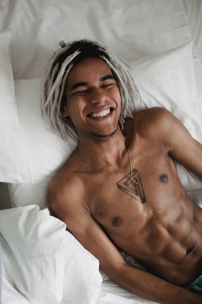 Ragazzo sorridente. Ritratto dell'uomo di colore africano bello con i dreadlocks che si trovano confortevolmente nel letto bianco a casa. Contrasto in bianco e nero