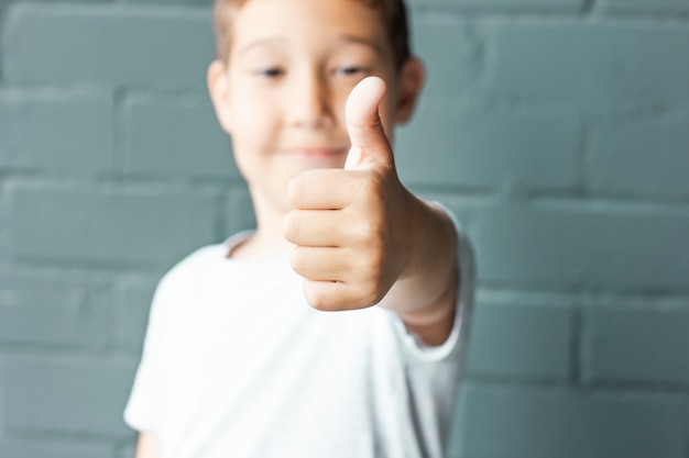 Ragazzo sorridente sveglio 8 anni che mostra i pollici su su fondo del muro di mattoni grigio