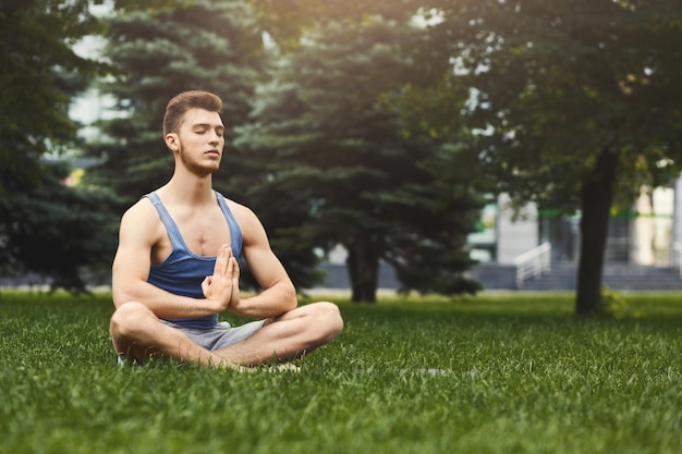 Ragazzo sportivo che pratica yoga seduto a padmasana. Giovane nella posa del loto, distogliendo lo sguardo, namaste su erba verde nel parco, copia spazio